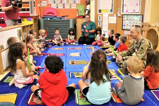 Young kids, a teacher and a garrison commander in uniform sit on the floor around a colorful rectangular rug.